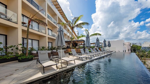 Zona junto a la piscina con sillones de descanso y sombrillas, un bar y un edificio con balcones.
