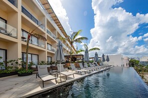 Zona junto a la piscina con sillones de descanso y sombrillas, un bar y un edificio con balcones.