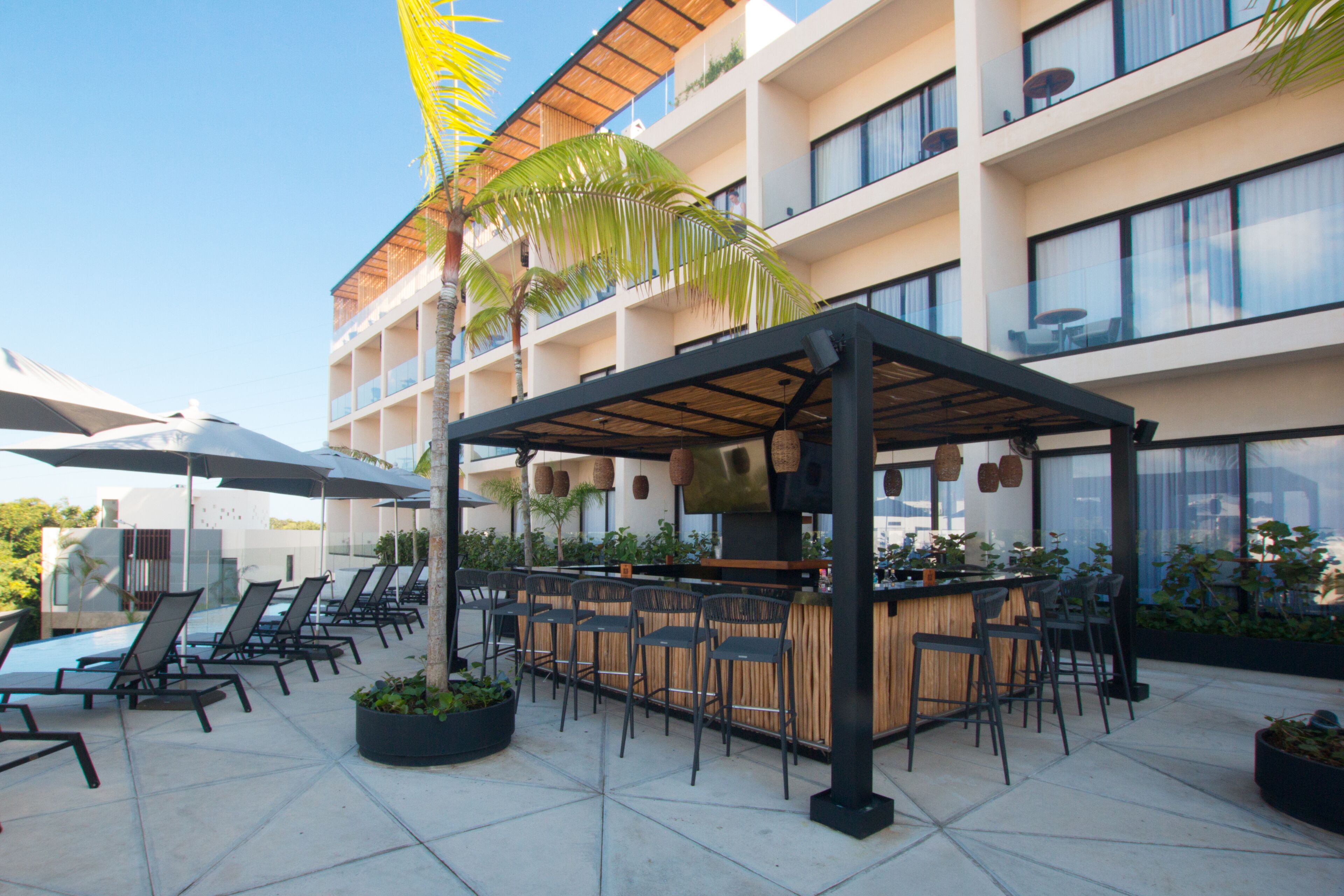 Un bar en la azotea con sillones de lounge y sombrillas, rodeado por un edificio moderno con balcones y ventanas amplias.