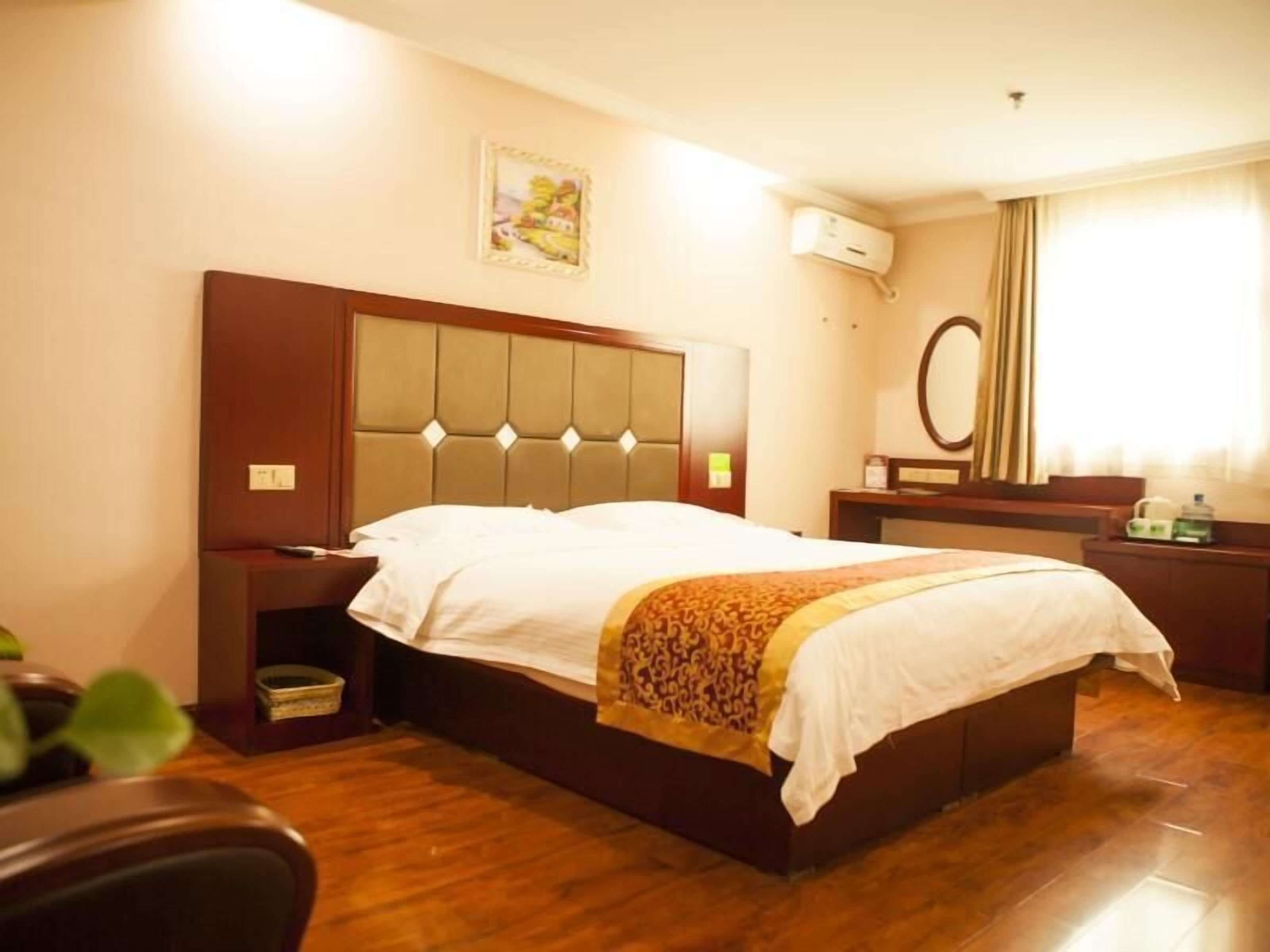 A hotel room with a large bed, a wooden headboard, a bedside table, a nightstand, a mirror, and a wall-mounted air conditioning unit.