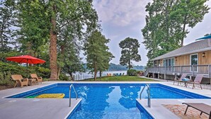 Une piscine rectangulaire entourée d’une terrasse en bois, avec un parasol rouge et des chaises longues.