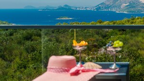 Een tafel met een roze hoed en twee fruitglazen op een balkon met uitzicht op een kustlandschap.
