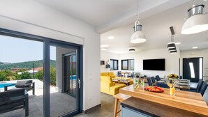 Een moderne keuken met een houten aanrecht, een eethoek met gele stoelen en een groot raam met uitzicht op een zwembad en groen.