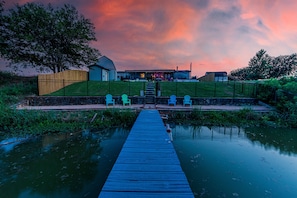 A wooden dock leading to a house with a porch, surrounded by greenery and a calm water body.
