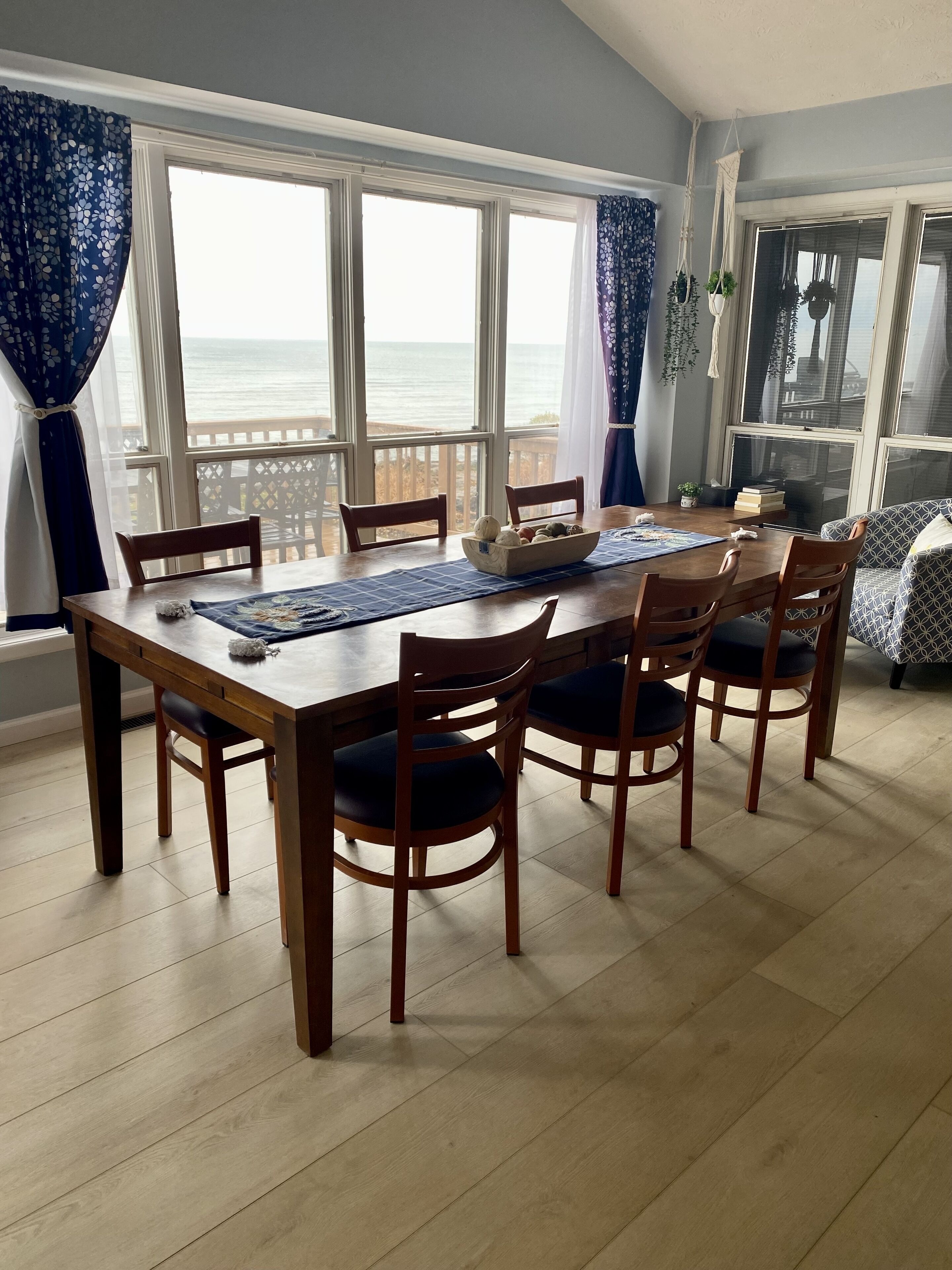 Un coin repas avec une table et des chaises en bois, de grandes fenêtres donnant sur la mer et un tapis à motifs.