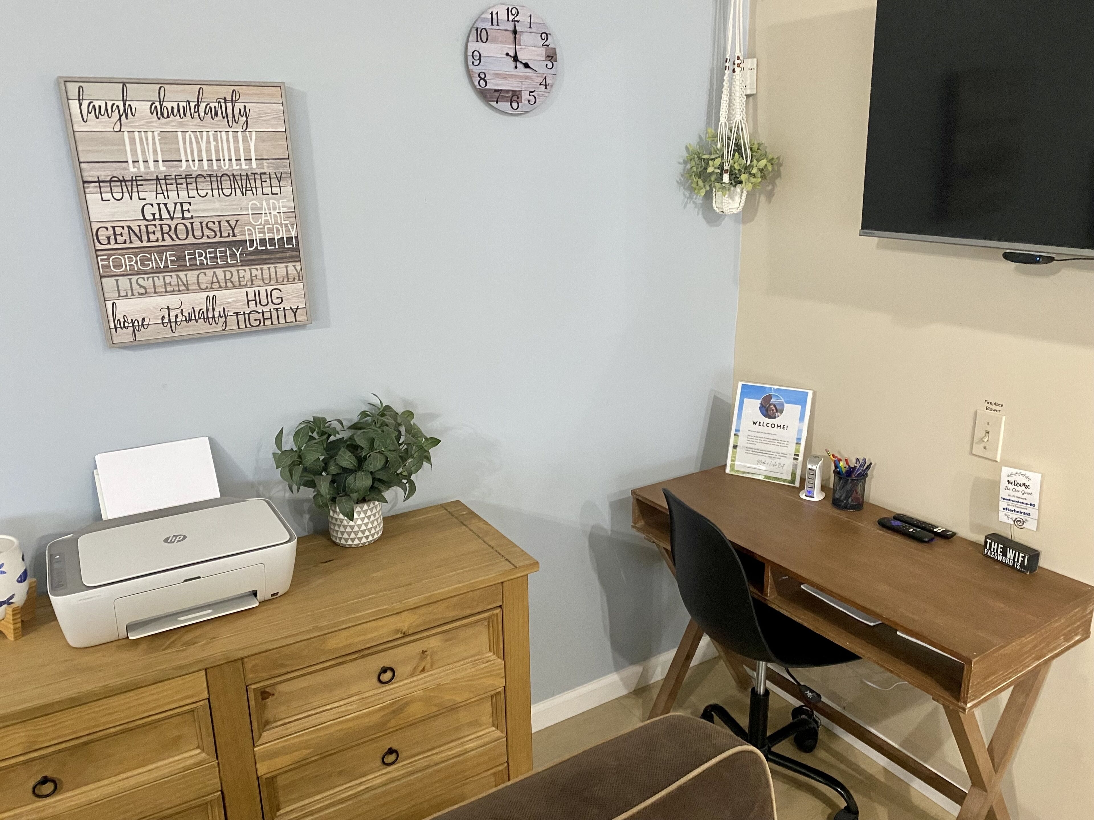 A home office with a wooden desk, a printer, a laptop, a clock, a welcome sign, and a plant.