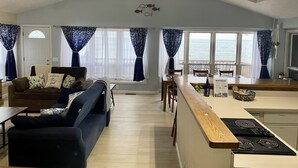 Amplia sala de estar con un sofá, comedor con mesa y sillas, cocina con estufa, y ventanas grandes con vista al mar.