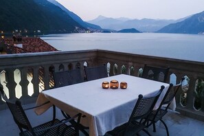 A table with a white tablecloth and three candles on a balcony overlooking a lake with mountains in the background.