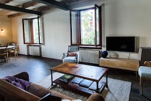 A cozy living room with a wooden coffee table, a sofa, a TV, and a window with a view of greenery.