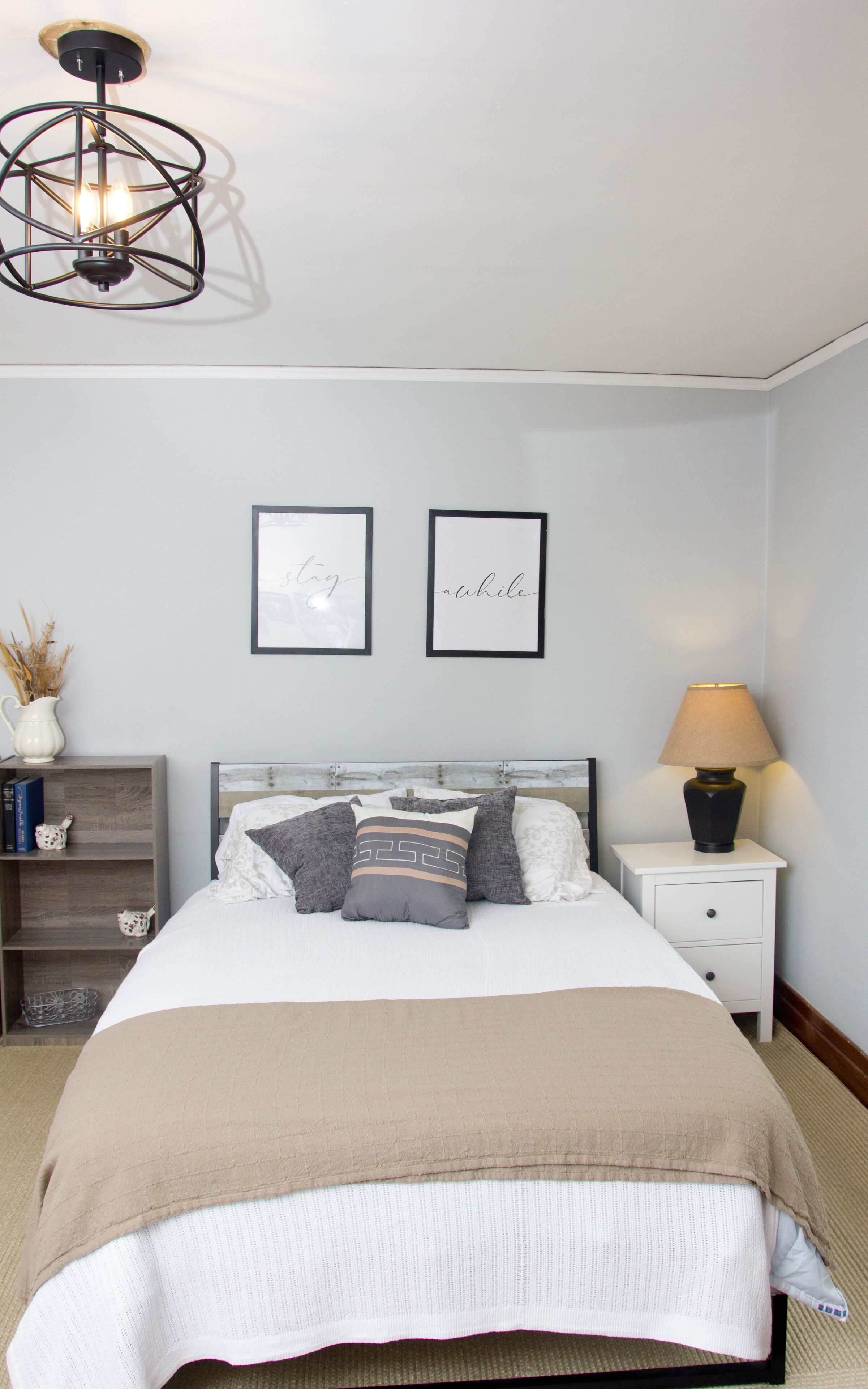 Un dormitorio con una cama, una mesita de noche con lámpara, un pequeño estante con libros y un objeto decorativo, y dos cuadros en la pared.