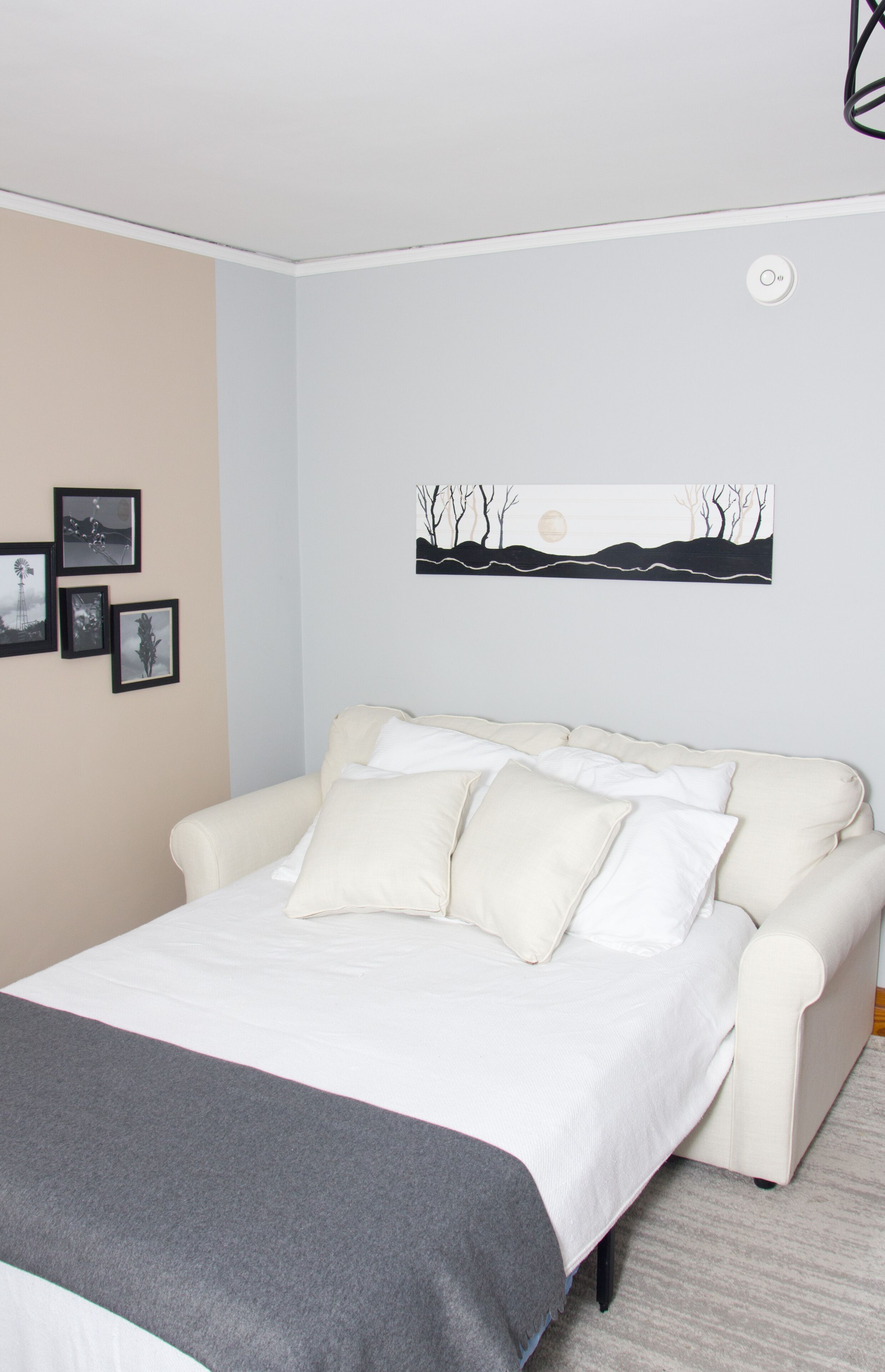 Une chambre avec un lit blanc, un plaid gris et un mur de couleur beige. Des tableaux encadrés ornent le mur, et une peinture est accrochée au-dessus du lit.