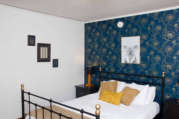 A neatly made bed with a headboard, a nightstand with a lamp, and a wall with a floral pattern and a framed picture.