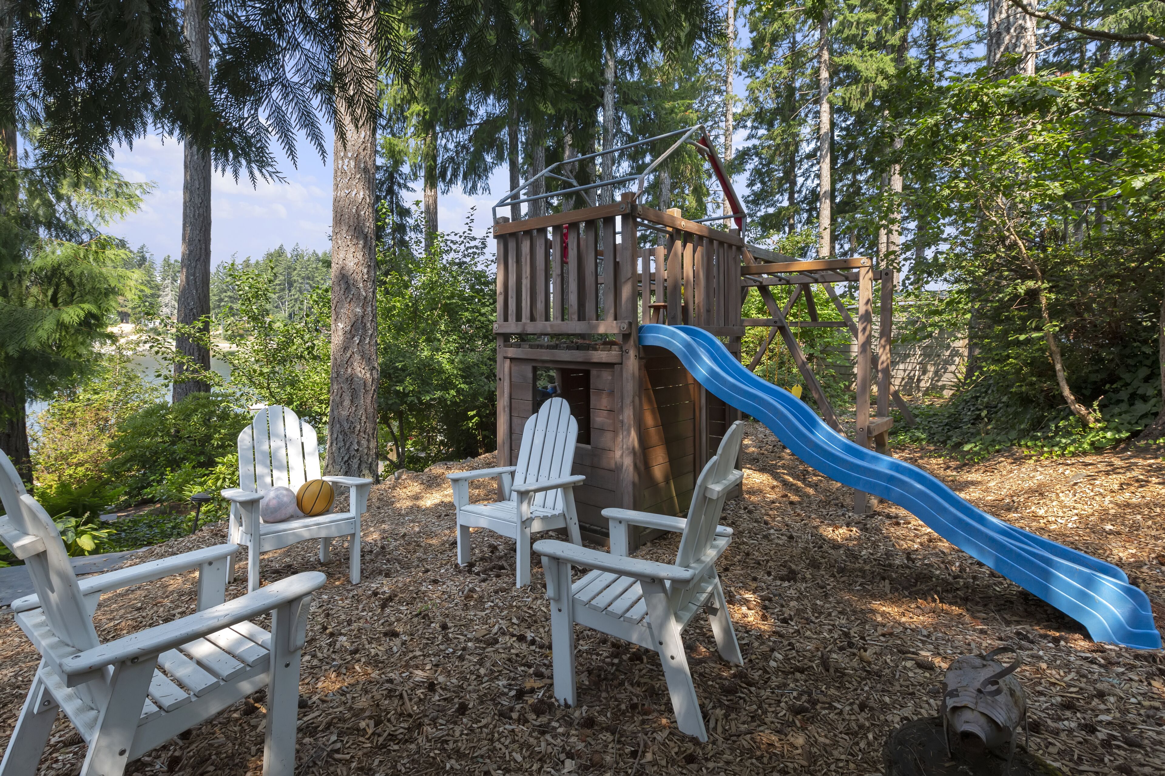 A wooden play structure with a slide, surrounded by trees and two Adirondack chairs.