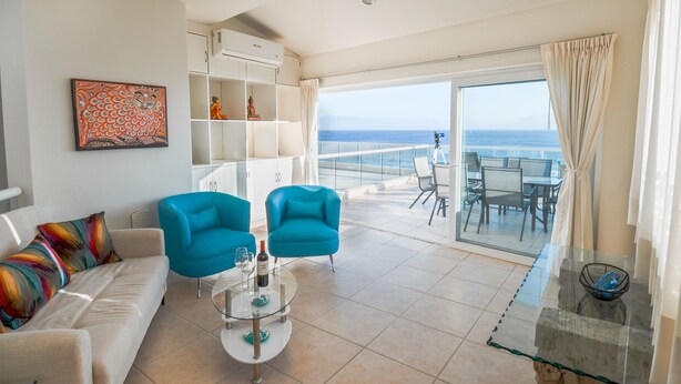 Een moderne woonkamer met een bank, twee blauwe fauteuils, een glazen salontafel en uitzicht op de oceaan door een schuifdeur.
