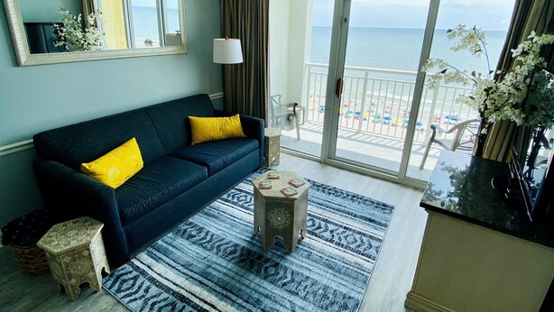 Un salon avec un canapé bleu, des coussins jaunes, un tapis à motifs, un petit tabouret en bois et une vue sur la plage par une porte vitrée.