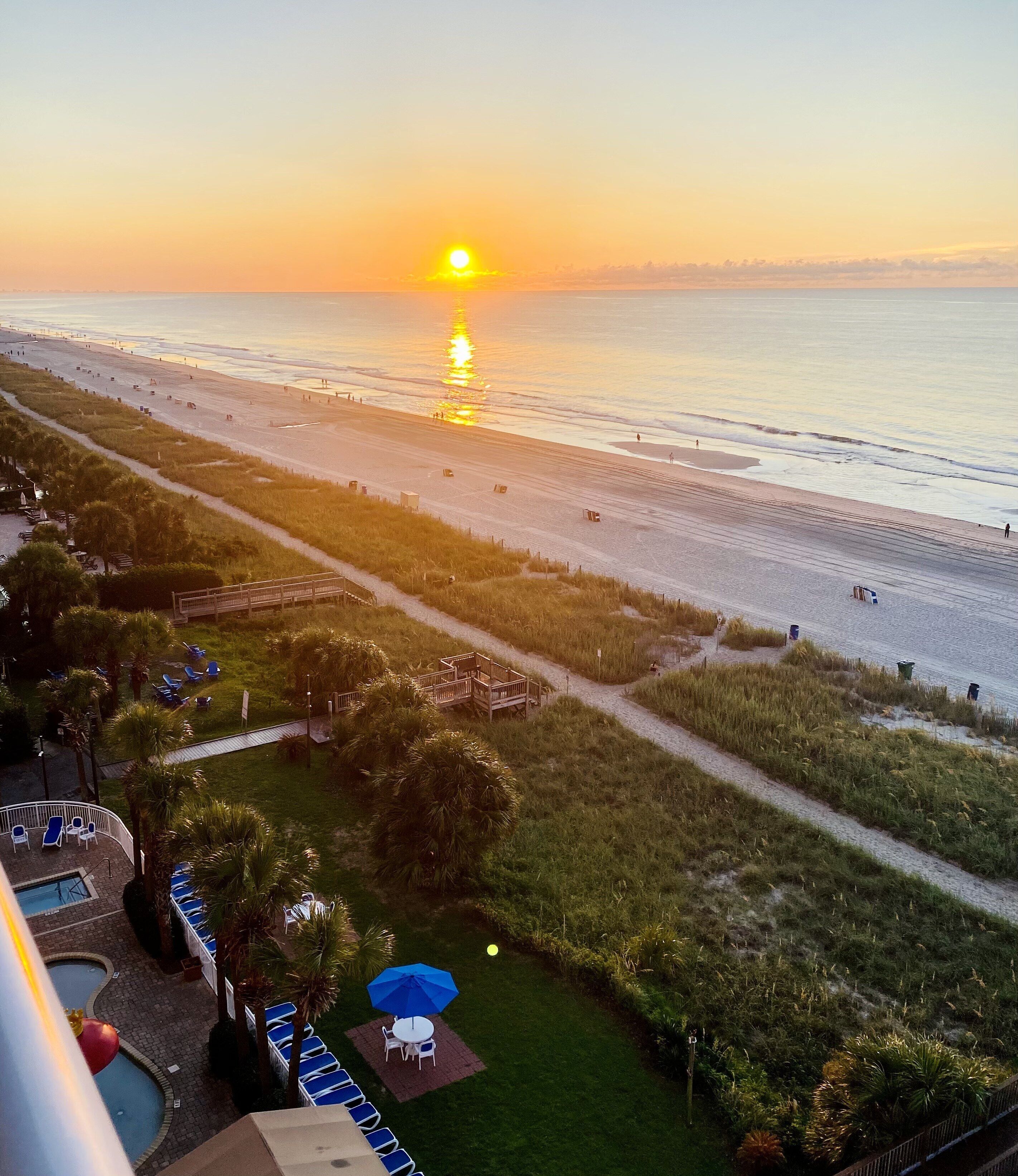 A beach view with a sunset, a pool area with lounge chairs, and a pathway leading to the beach.