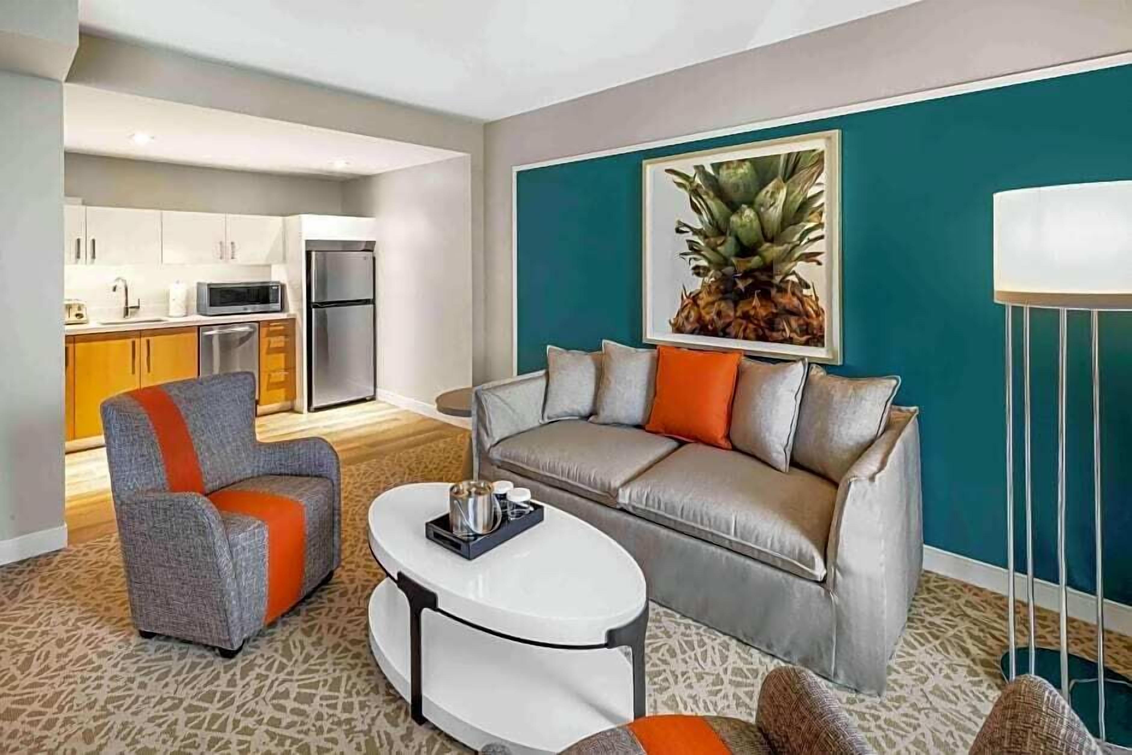 A modern living room with a grey sofa, orange and grey armchair, and a white coffee table. A kitchen is visible in the background with white cabinets and a microwave.