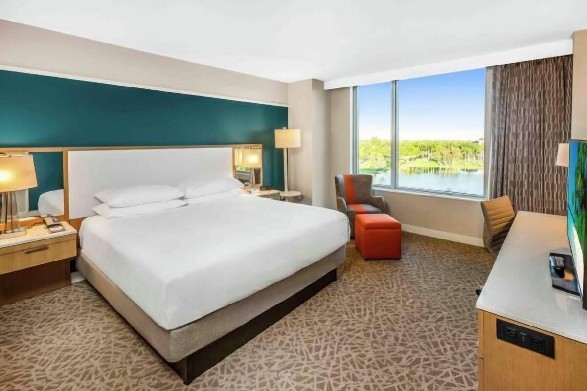Ein Hotelzimmer mit einem großen Bett, einem Schreibtisch, einem Sessel und Blick auf einen See.