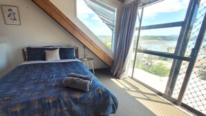A bedroom with a large bed, a small table, and a view of a river through large windows. - Relaxing Spot with Stunning Sea View! (Snells Beach)