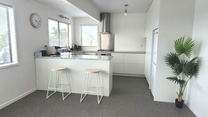 A modern kitchen with white cabinetry, a central island with stools, a clock, and a potted plant. - Relaxing Spot with Stunning Sea View! (Snells Beach)