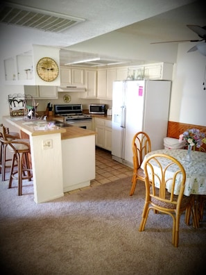 A small kitchen with white cabinets, a microwave, a stove, and a refrigerator. There is a dining table with chairs and a clock on the wall.