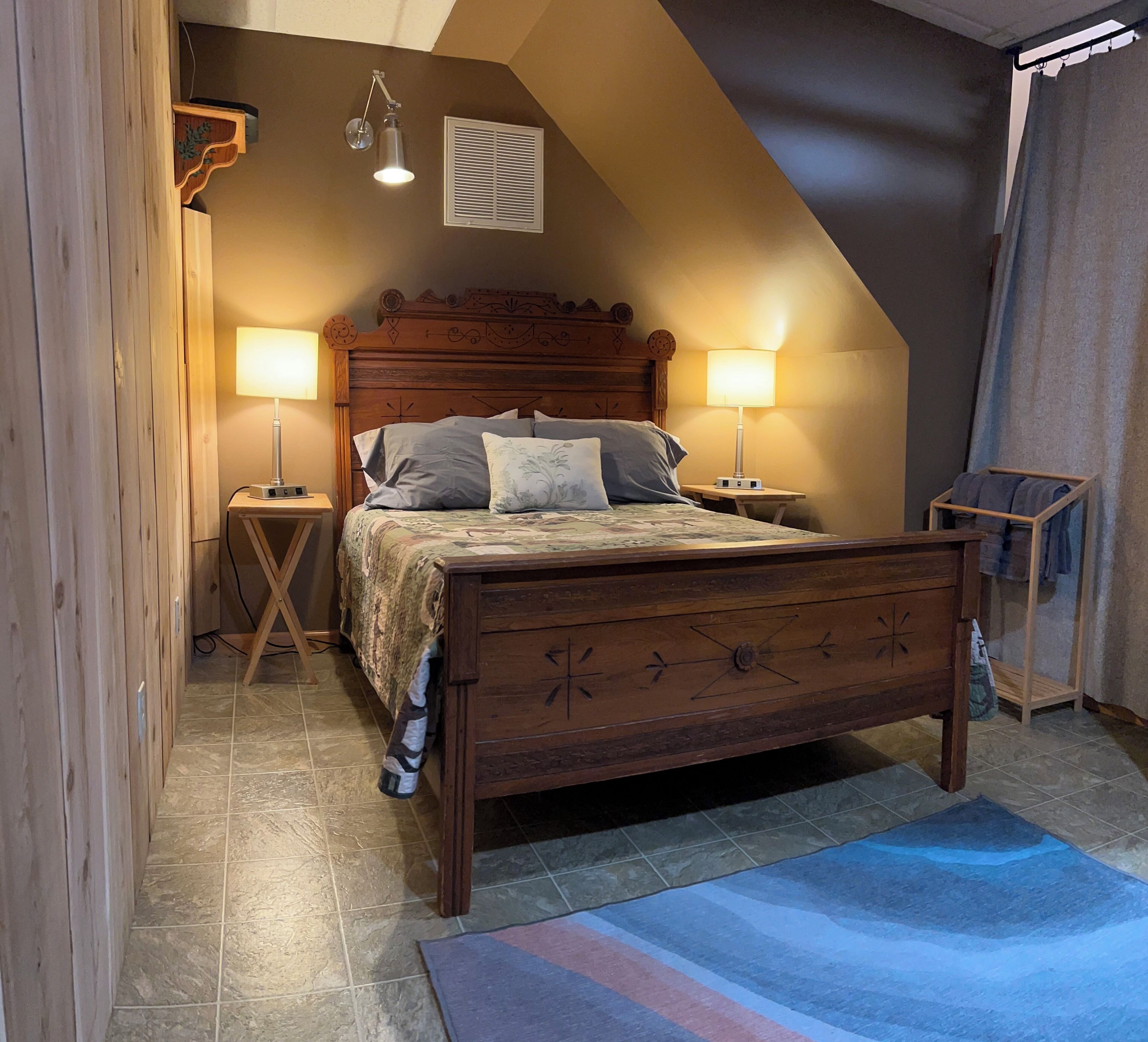A bedroom with a wooden bed, two bedside lamps, a nightstand, a wooden wardrobe, a window with blinds, and a blue rug.