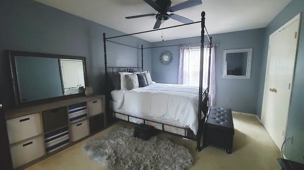 A bedroom with a four-poster bed, a ceiling fan, a flat-screen TV, a clock, and a window with curtains.