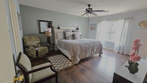 Quarto com cama, cadeira, escrivaninha, ventoinha no teto, janela com cortinas e relógio na parede.