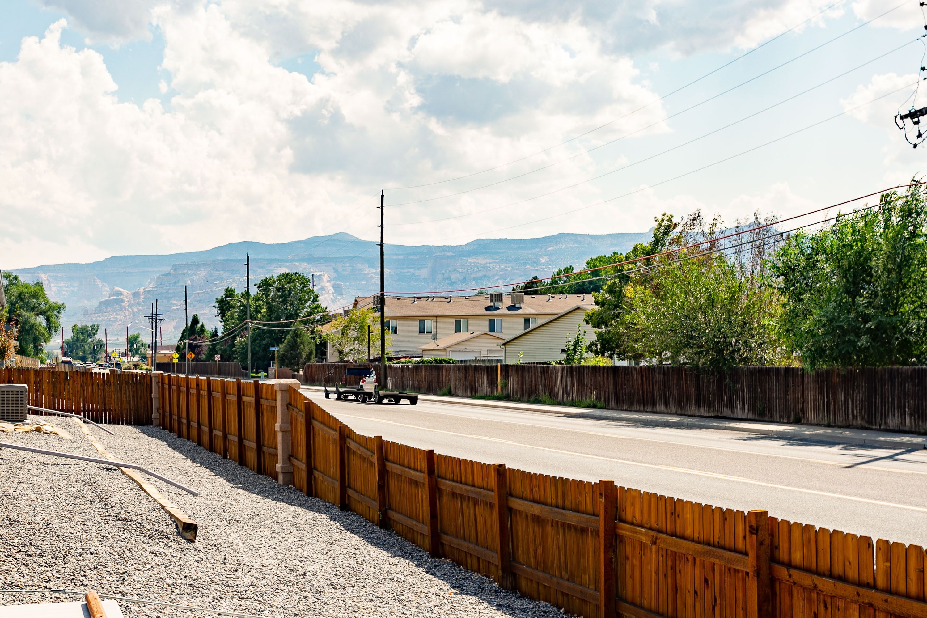 A road with a wooden fence on one side and a residential area with houses and mountains in the background.