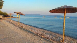 A beach with pebbles and a clear blue sea. There are umbrellas and a person walking. - Villa in Malaki in Sea With PlayGround (Malaki)