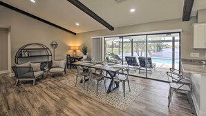Uma sala de estar ampla com uma mesa de jantar, cadeiras e um sofá. Há uma grande janela com vista para uma piscina e área de estar ao ar livre.