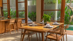 Un comedor con mesa y sillas de madera, ventanas amplias con vistas a zonas verdes y suelo de hormigón.