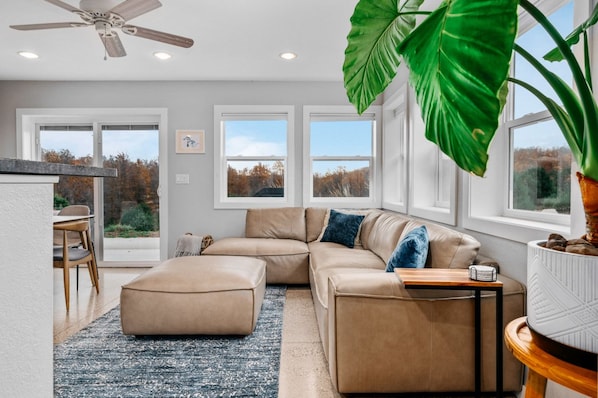 A modern living room with a sectional sofa, a dining area with a table and chairs, large windows with a view of trees, and a ceiling fan.
