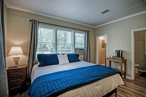 Une chambre avec un grand lit, une table de chevet avec une lampe, une petite table avec une machine à café et une porte menant à la salle de bain.