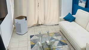 Uma sala de estar com um sofá branco, uma mesa de centro de vidro, um tapete estampado, uma televisão e um quadro fixado na parede.
