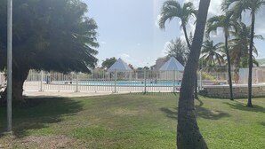 Piscina cercada por uma cerca branca, com palmeiras e uma área gramada ao primeiro plano.