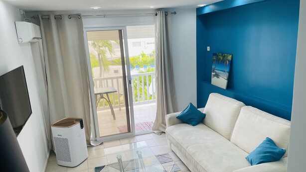 Uma sala de estar com um sofá branco, uma mesa de centro de vidro, uma televisão e uma porta de correr que dá para uma varanda.