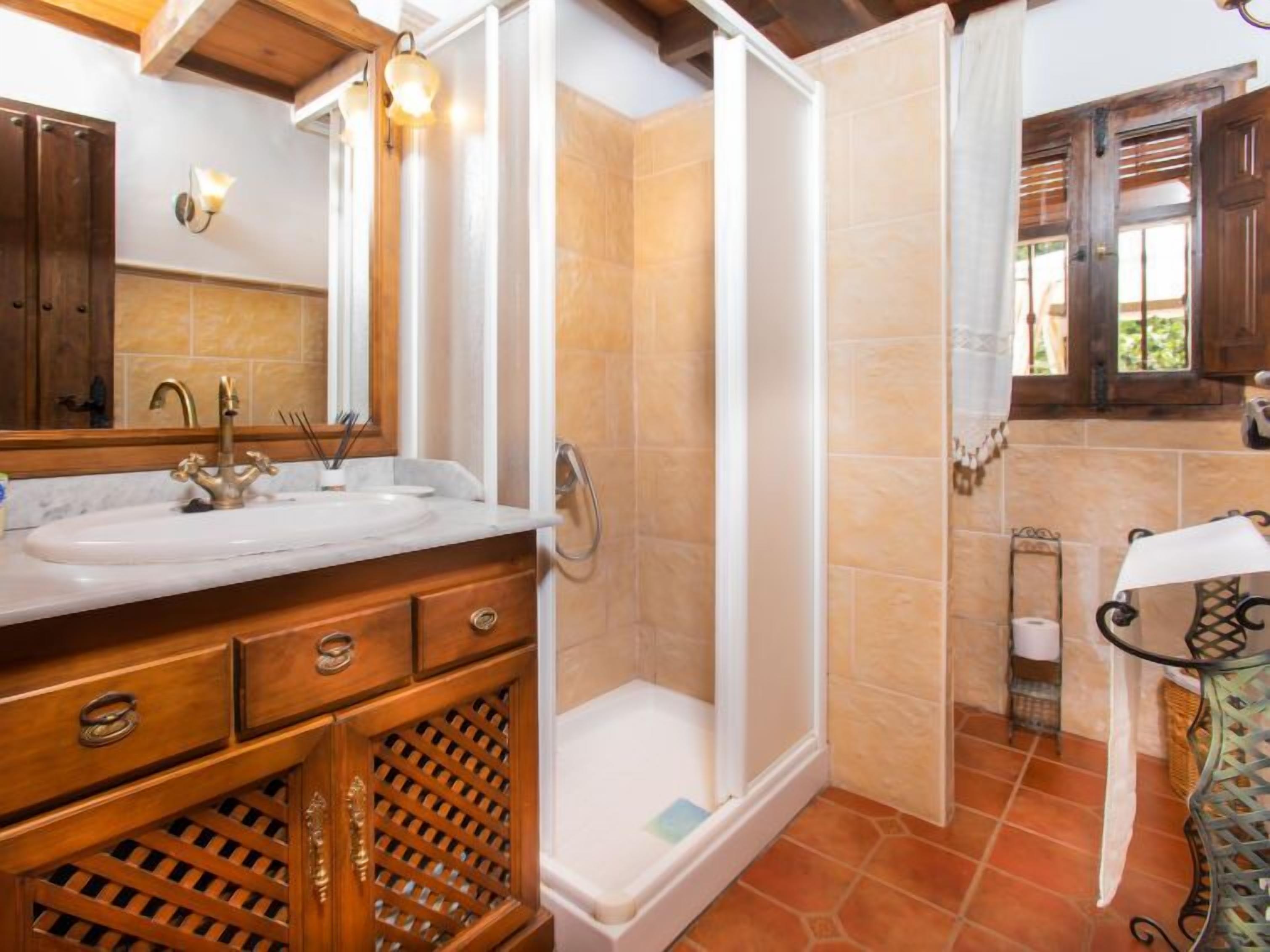 A bathroom with a marble sink, wooden vanity, a shower enclosure, and a window with a view of greenery.