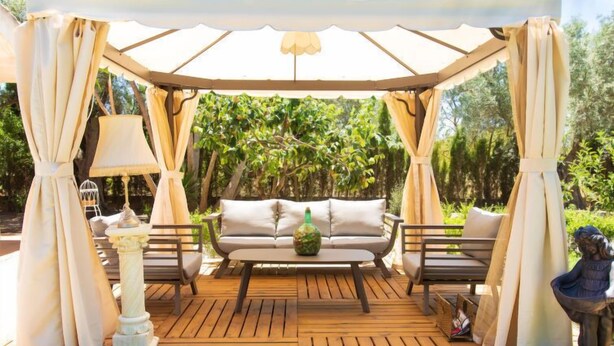 Une terrasse en bois dotée d’un espace salon couvert, comprenant un canapé et deux fauteuils, le tout entouré de rideaux et agrémenté d’une lampe.