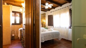 Une chambre à coucher comprenant un lit, une fenêtre avec des rideaux, un ventilateur de plafond et une salle de bain avec une douche et un porte-serviettes.