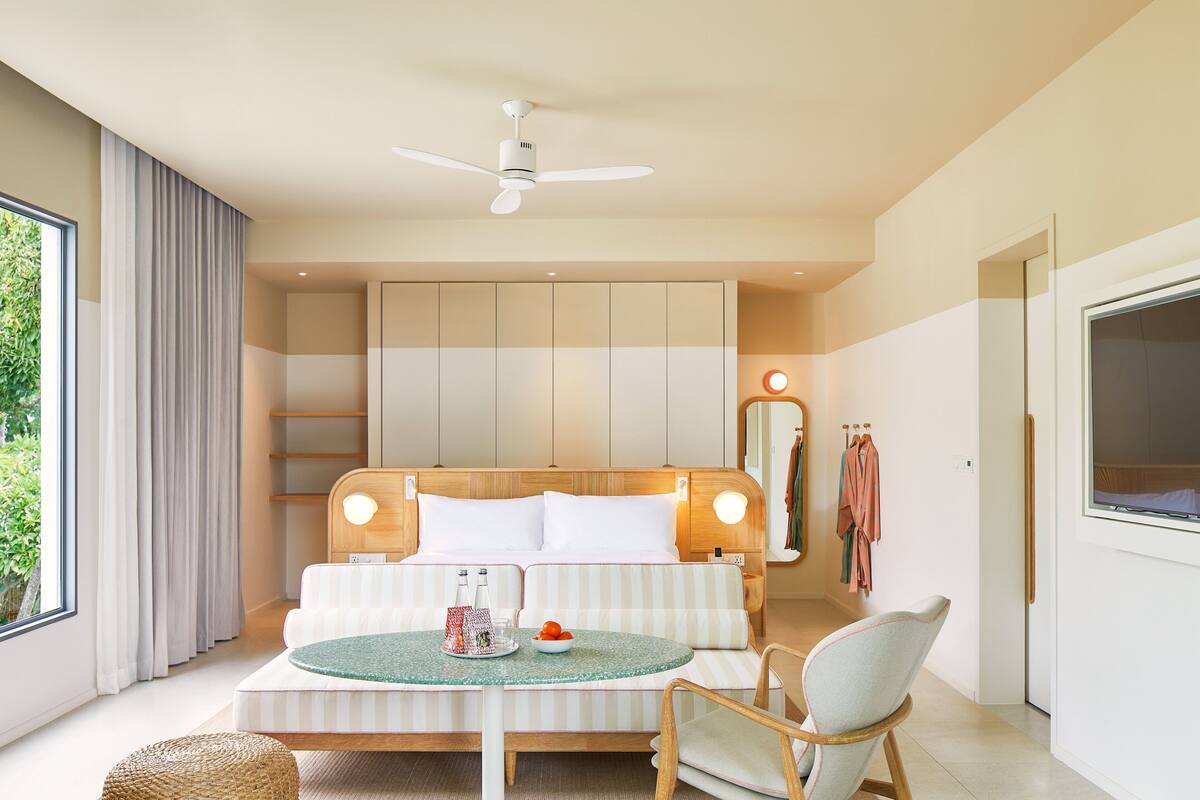 Ein modernes Hotelzimmer mit einem großen Bett, einem runden Esstisch, einem Stuhl, einem Deckenventilator und einem großen Fenster.