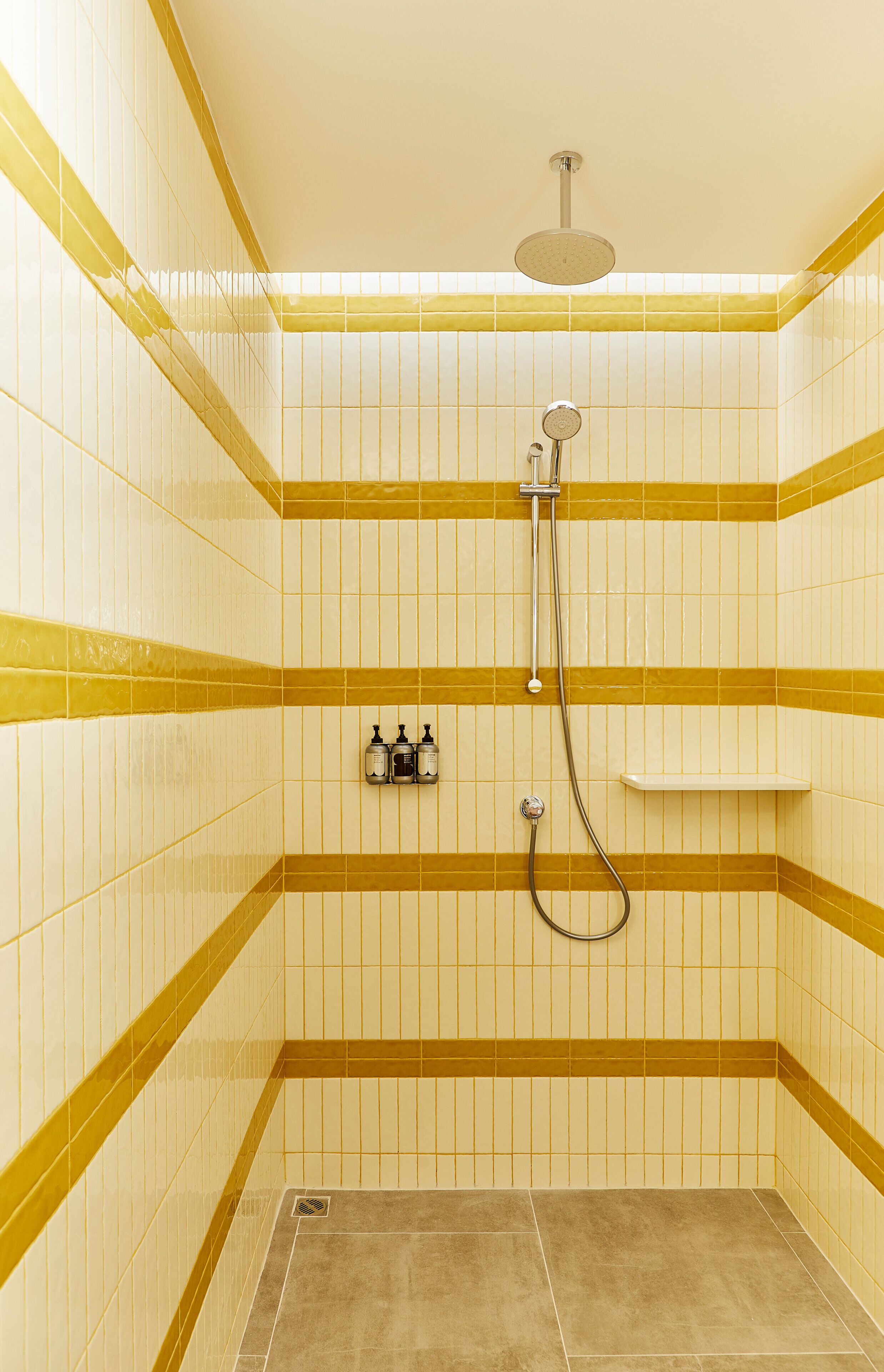 Un área de ducha con azulejos blancos y amarillos, un cabezal de ducha manual y un grifo instalado en la pared.