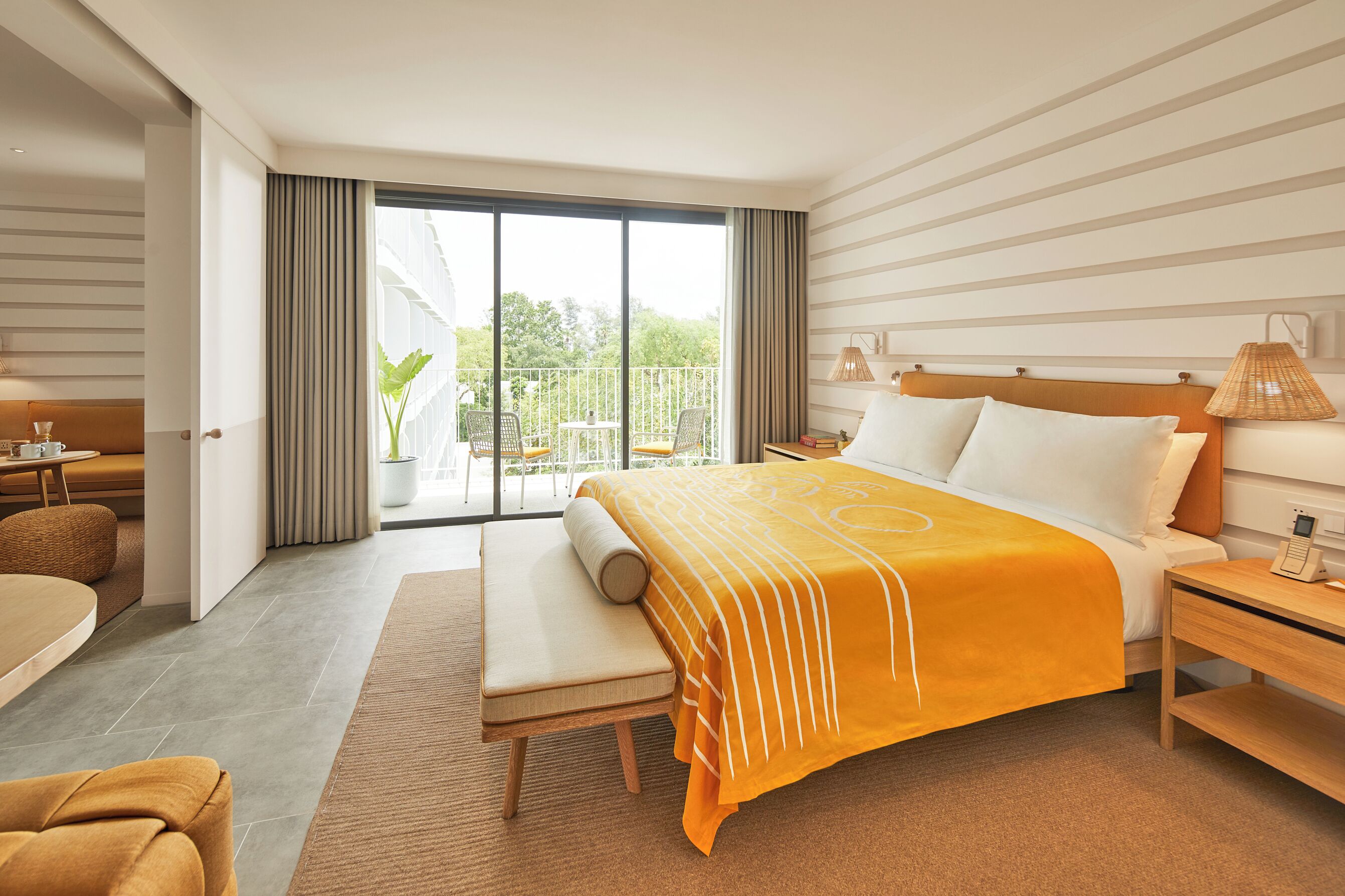 A modern hotel room with a large bed, a bedside table, a chair, a sofa, and a balcony with outdoor seating.