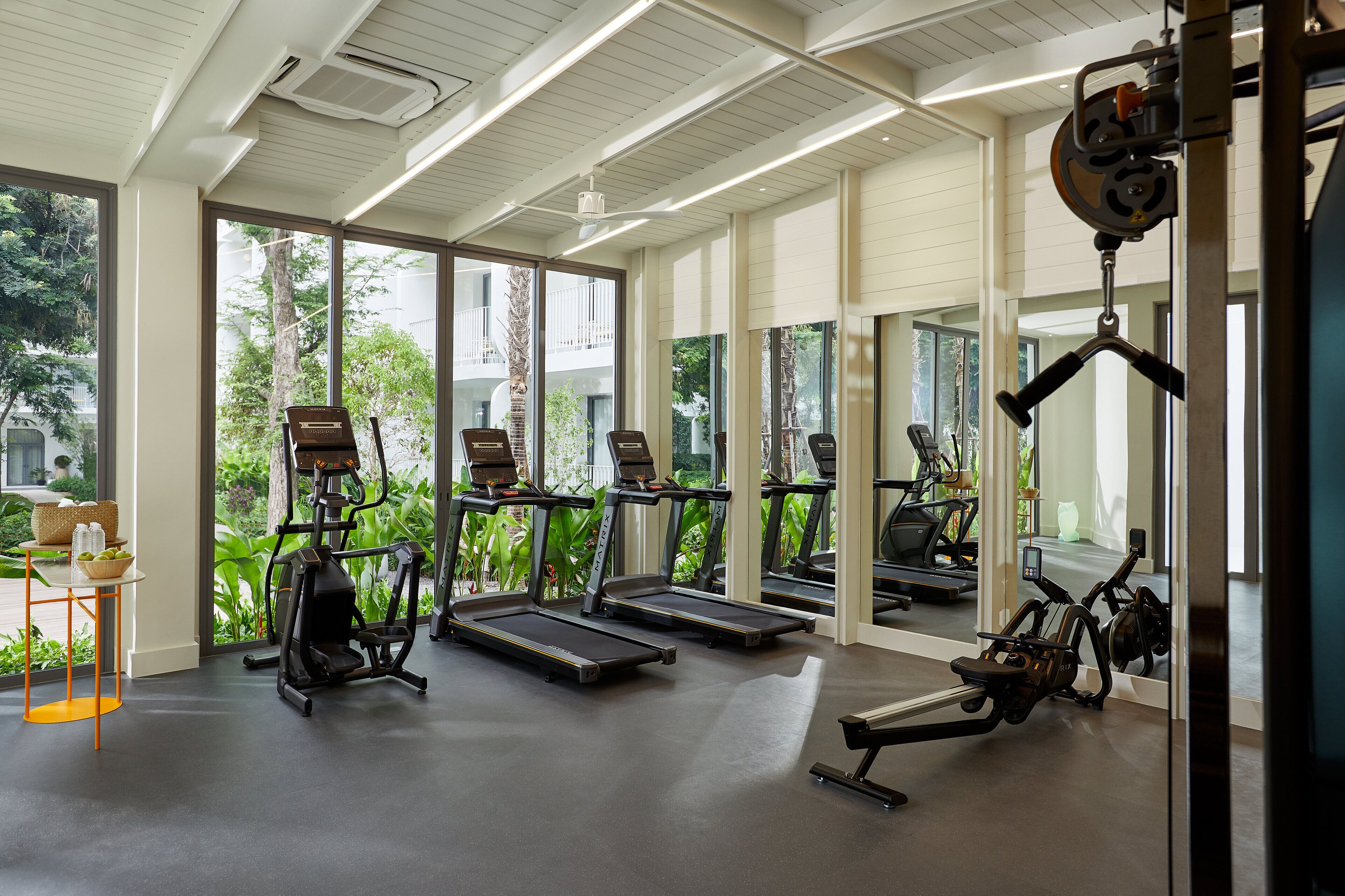 A modern gym with treadmills, elliptical machines, and a rowing machine. Large windows offer views of greenery and buildings outside.