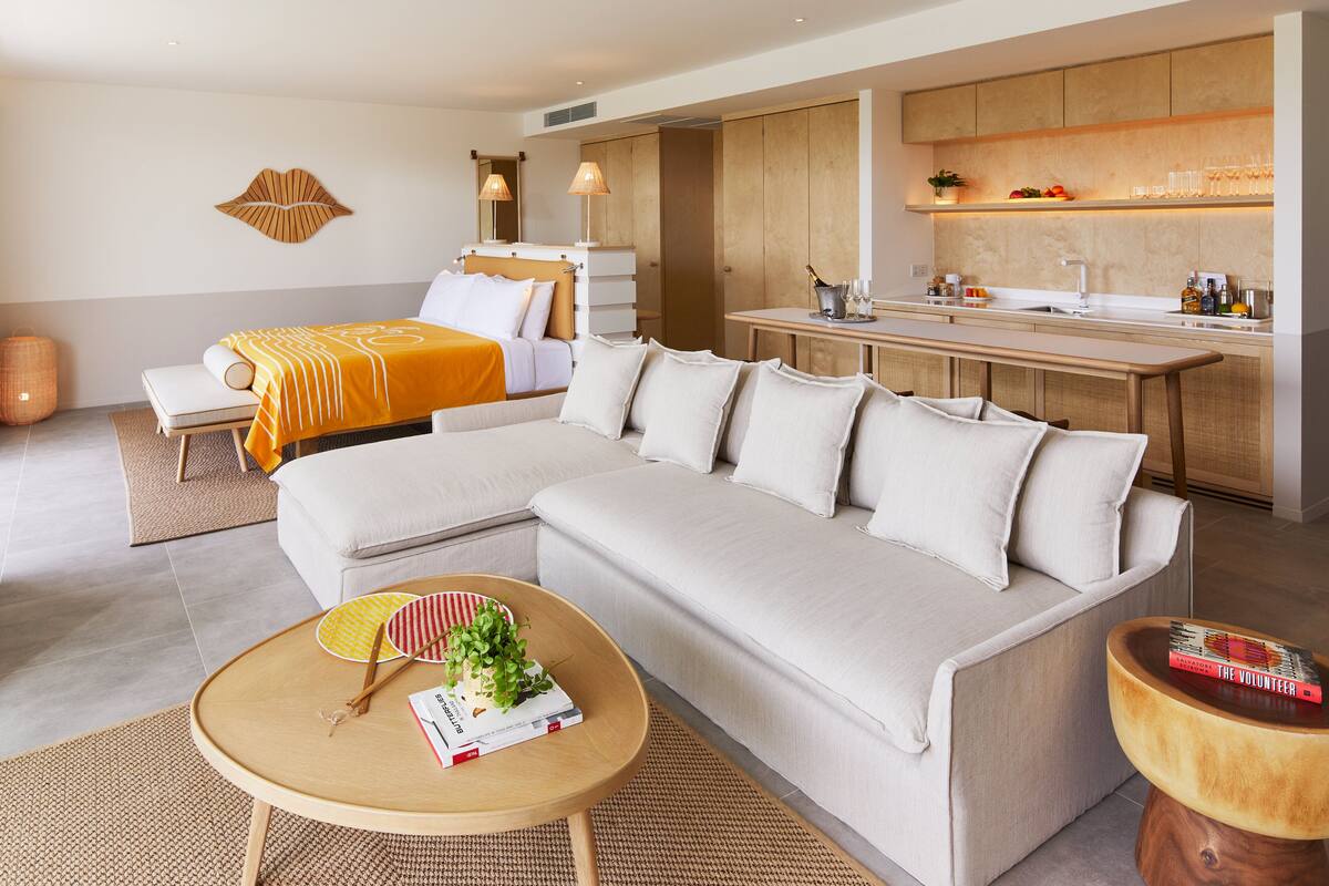 Ein modernes Hotelzimmer mit einer großen Couch, einem Esstisch, einer kleinen Küchenzeile und einem Bett mit gelber Tagesdecke.