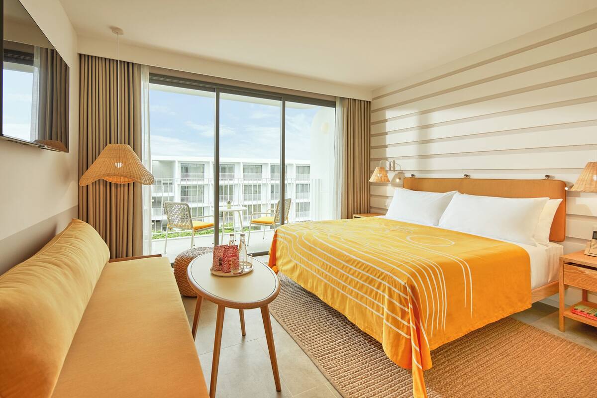 Ein Hotelzimmer mit Bett, Sofa, kleinem Tisch und Blick auf ein Gebäude durch große Fenster.