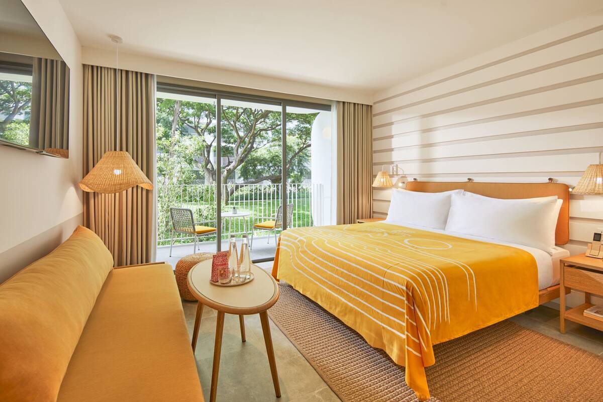 A hotel room with a bed, a sofa, a small table with a lamp, and a view of greenery outside.