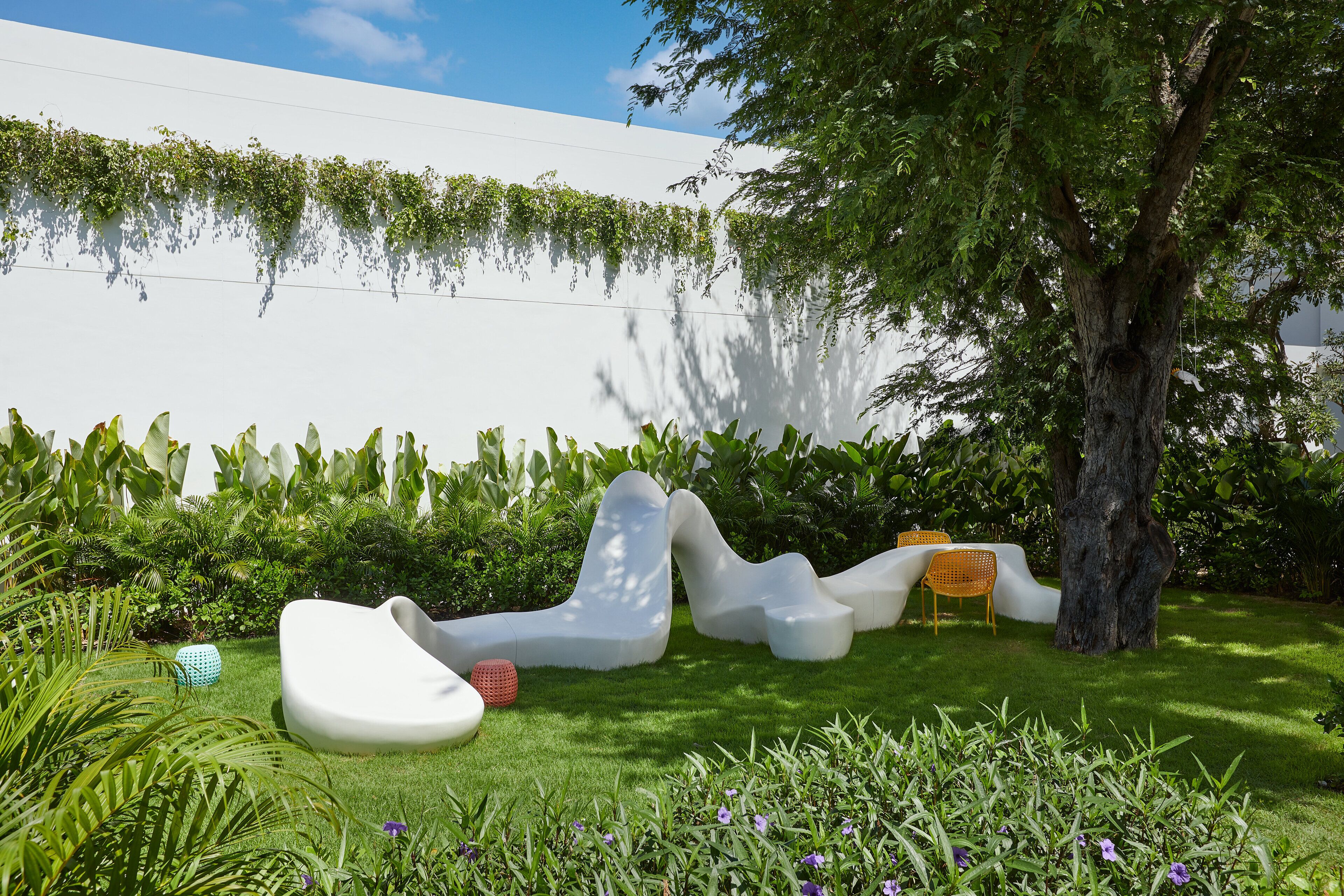 A modern outdoor seating area with white, sculptural furniture, surrounded by lush greenery and a tree.