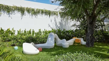 A modern outdoor seating area with white, sculptural furniture, surrounded by lush greenery and a tree.