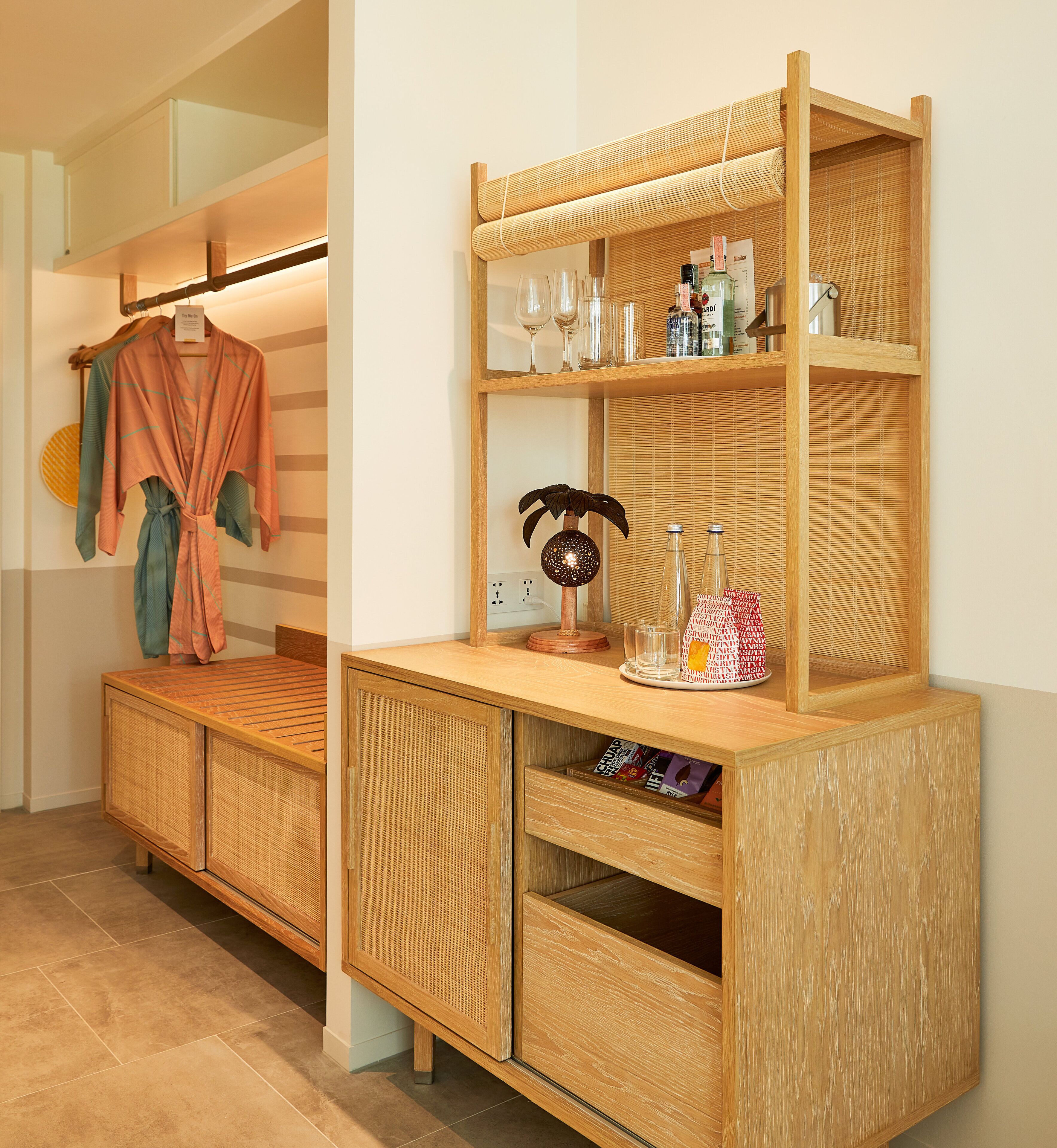 A wooden bench with a woven backrest, a shelf with bottles and glasses, and a hanging robe.